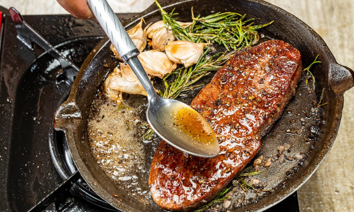 A person is cooking a sirloin steak in a pan with herbs and spices. The steak is brown and he is well-cooked