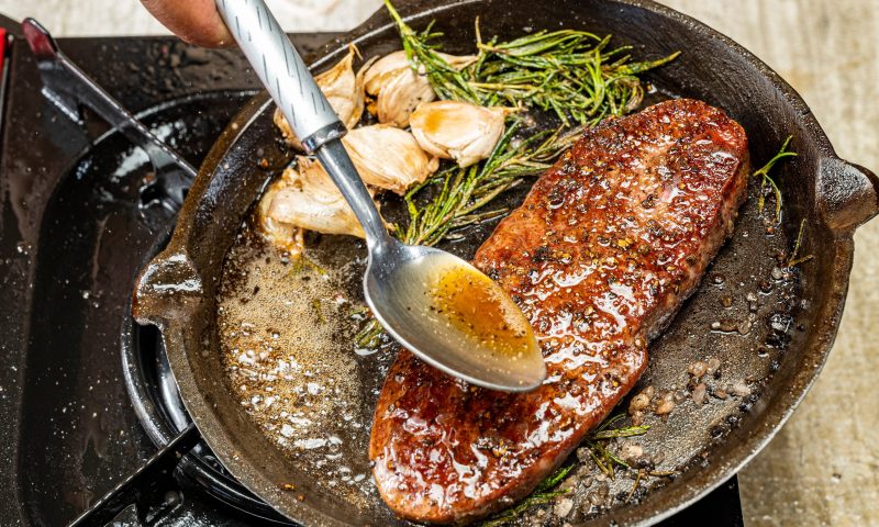 A person is cooking a sirloin steak in a pan with herbs and spices. The steak is brown and he is well-cooked