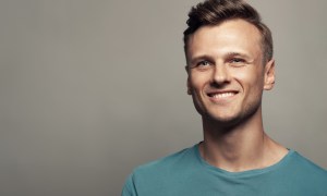Male beauty concept. Portrait of smiling 30-year-old man standing in blue t-shirt over gray background . Close up. Classic style. Wavy glossy blond hair. Studio shot