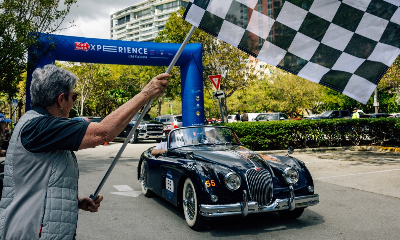 A Jaguar XK150 S crossing the finish line at the 1000 Miglia Experience Florida with a checkered flag waving