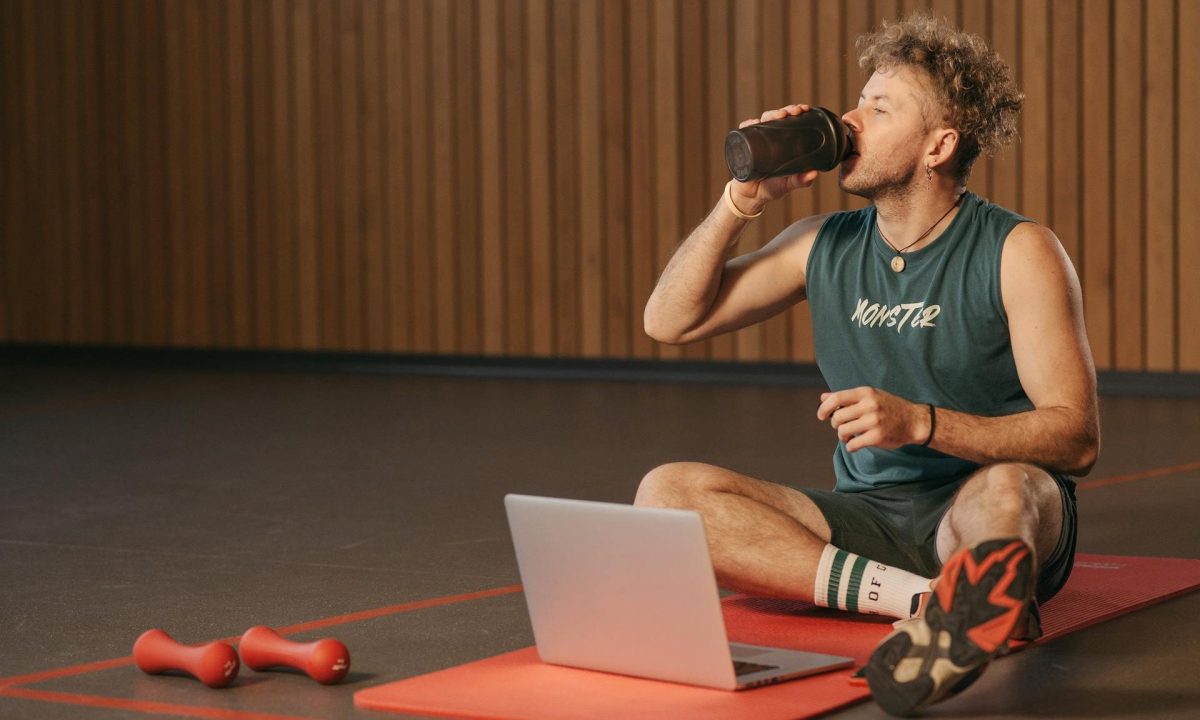 man drinking protein shake on a yoga mat with a laptop
