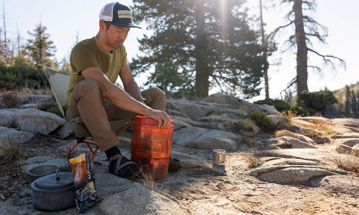 Young man camping in the woods with an REI Traverse bear-proof canister.