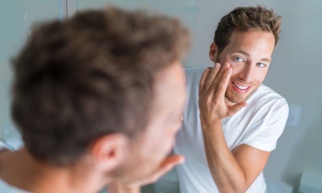 Man putting skincare facial treatment cream on face. Anti-aging skin care product. Male beauty morning routine at home lifestyle. Guy looking in bathroom mirror applying moisturizer under eyes.