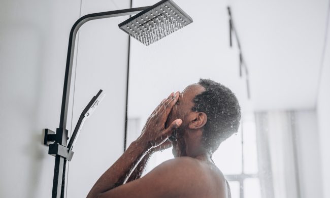 Relaxed young African-American man takes shower standing under hot water jets in contemporary unit in bathroom close side view