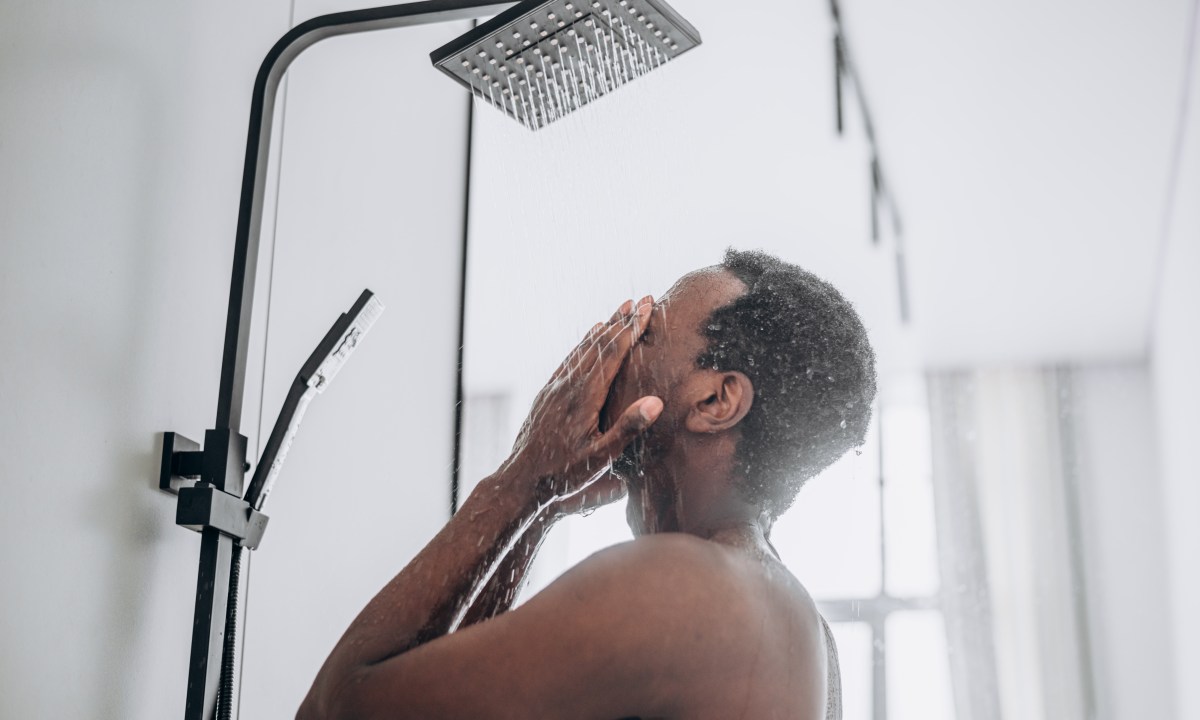 Relaxed young African-American man takes shower standing under hot water jets in contemporary unit in bathroom close side view