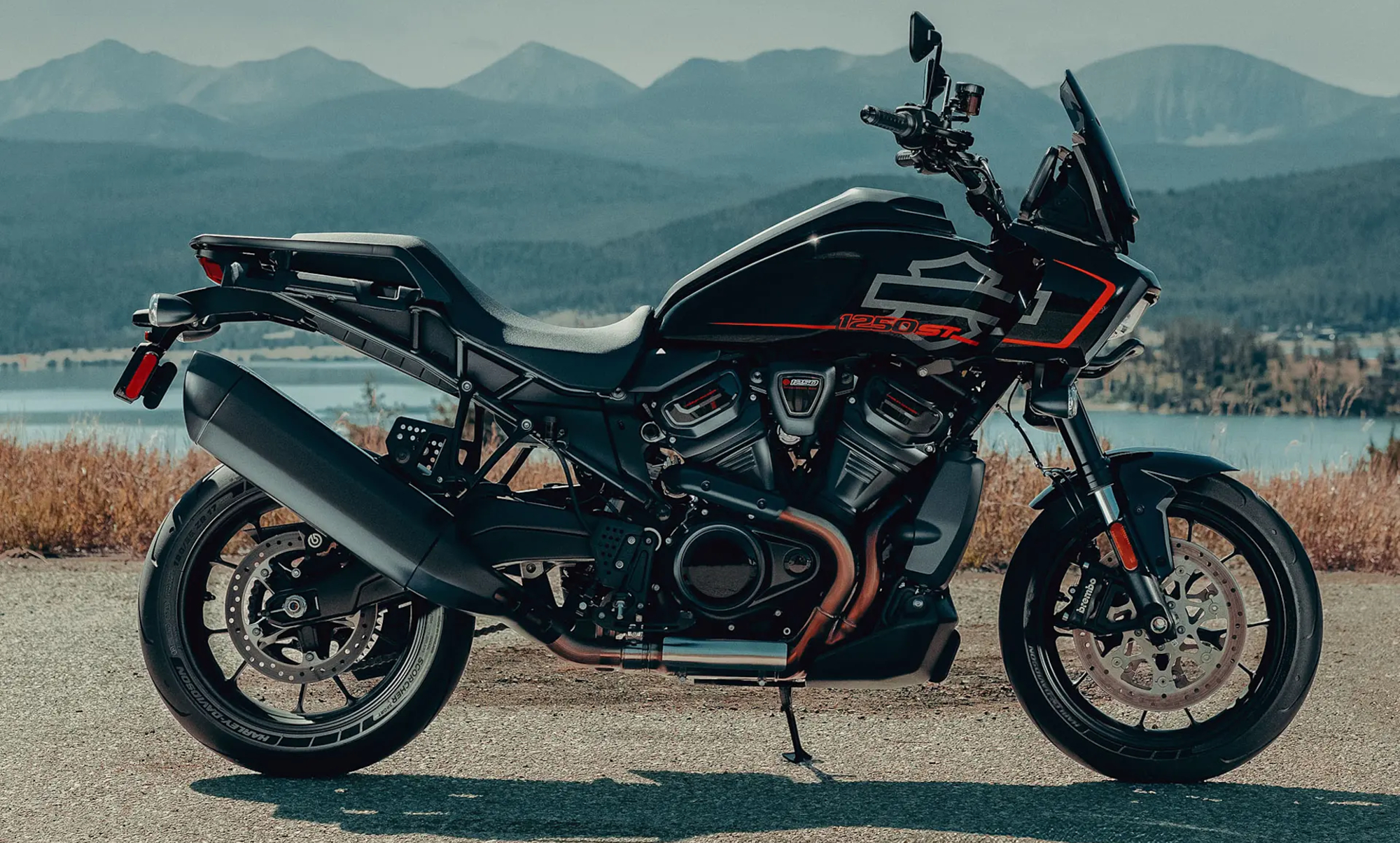 2025 Harley Davidson Pan America 1250 ST parked on pavement with a lake and mountains in the background.
