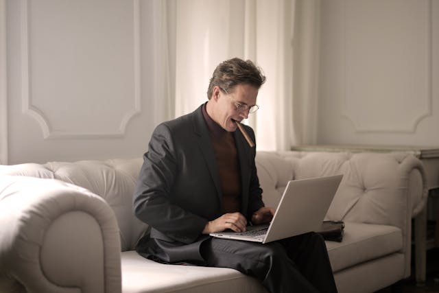 man in black suit sitting on white couch smoking a cigar while using his laptop