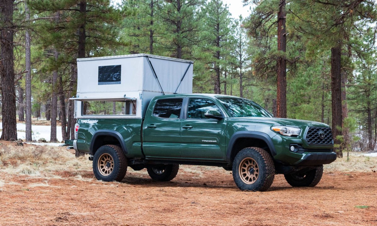 A green Toyota pickup truck parked in the woods with an Ovrlnd Campers Bivy Chubby truck camper mounted to the bed.