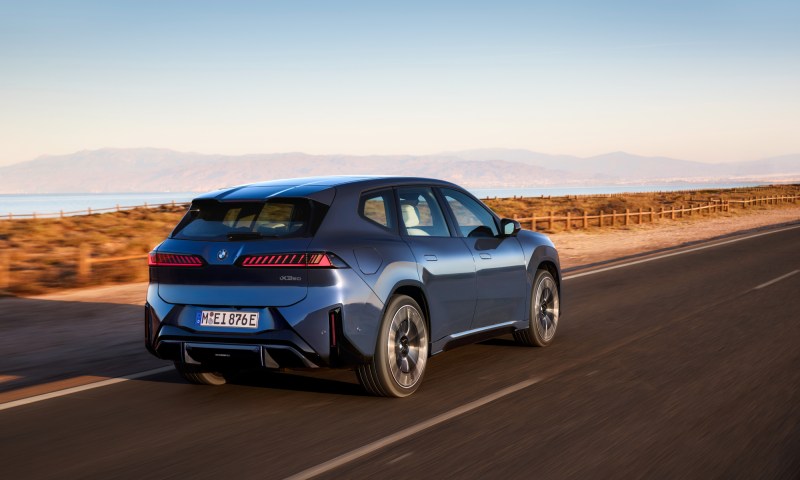 2026 BMW iX3 50 exterior right rear three quarter view driving on a highway with mountains in the far distance.