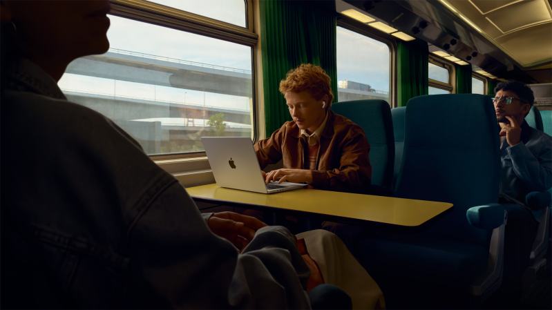 Man on a train working on a MacBook while wearing AirPods Pro 3 earbuds