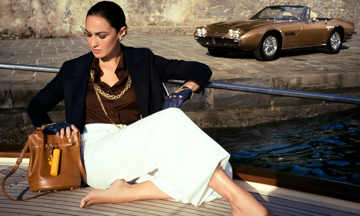 Model sitting on the deck of a yacht with The Art of Travel Kit and a Maserati Ghibli Spyder parked on shore.